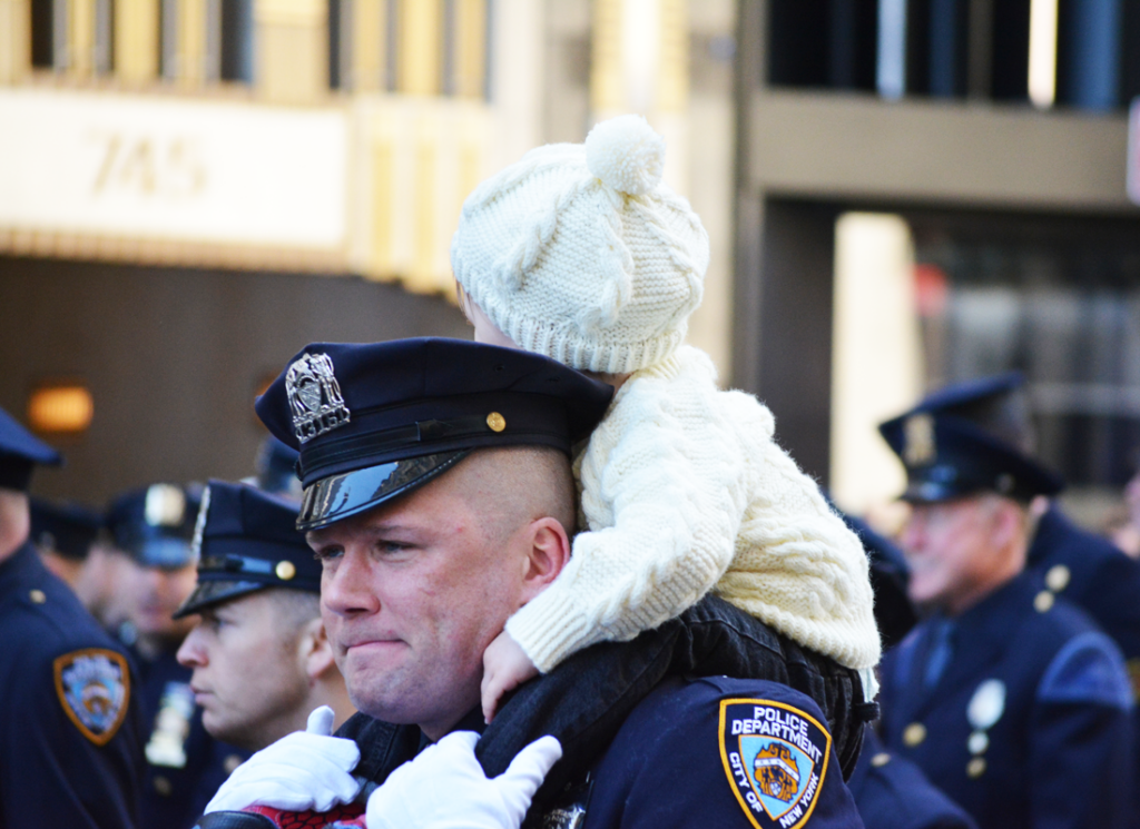Police officer carrying baby in white knit sweater and hat on shoulders